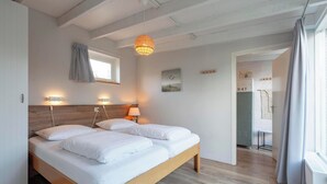 A modern bedroom with two single beds, a wooden headboard, bedside lamps, a window with curtains, and a view of a hallway with a door. - Duinland 247modern Bungalow for 6 Guests (Sint Maartensvlotbrug)