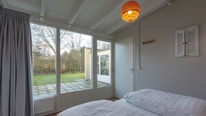 A bedroom with a large sliding glass door leading to a patio, a bed with white bedding, a hanging light fixture, and a white door with shutters. - Duinland 247modern Bungalow for 6 Guests (Sint Maartensvlotbrug)