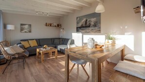 A modern living room with a wooden dining table, a sofa, and a chair. There is a painting on the wall and a window providing natural light. - Duinland 247modern Bungalow for 6 Guests (Sint Maartensvlotbrug)