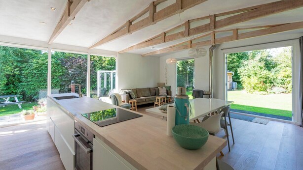 Une cuisine moderne avec un îlot central, un coin repas avec une table et des chaises, et une vue sur un jardin grâce à de grandes fenêtres.