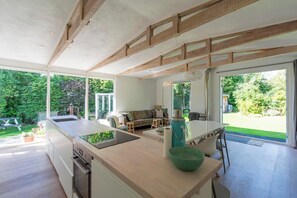 Een moderne keuken met een kookeiland, een eethoek met een tafel en stoelen, en een uitzicht op een tuin door grote ramen.