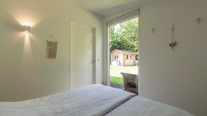 Une chambre avec un lit, une lampe fixée au mur, une décoration murale en macramé et une vue sur une maison par une porte ouverte.