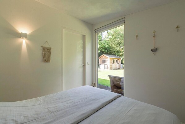 A bedroom with a bed, a wall-mounted light, a macrame wall hanging, and a view of a house through an open door. - Eureka 109 Bungalow for 6-person With Garden (Sint Maartensvlotbrug)