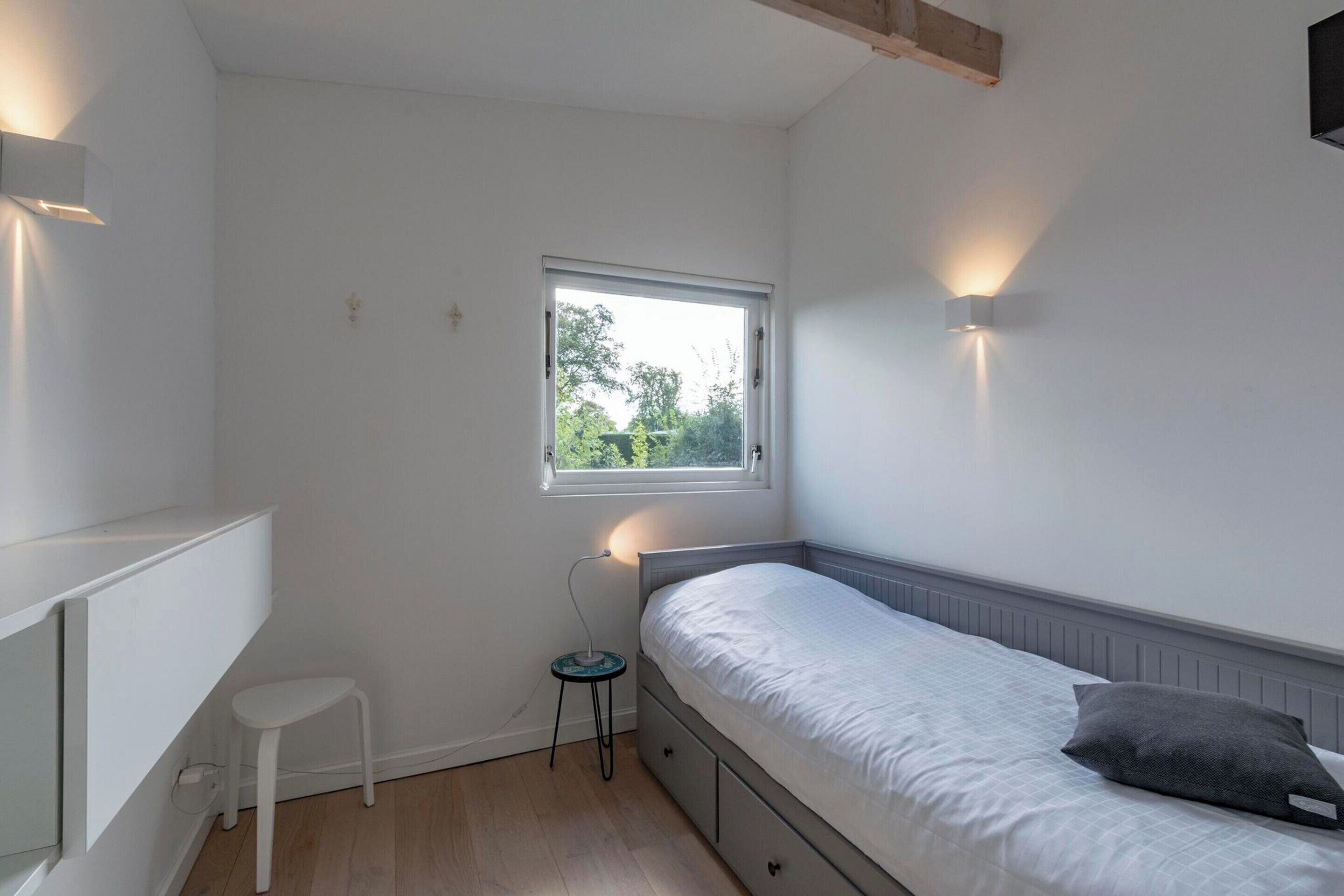 Ein kleines, helles Schlafzimmer mit einem Einzelbett, einem kleinen Hocker und einem Fenster, das natürliches Licht hereinlässt.
