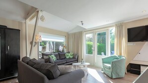 A modern living room with a sofa, a small table, and a chair. There is a television mounted on the wall and a window with curtains. - Holiday Home Sint Maartenszee Near Beach (Sint Maartensvlotbrug)