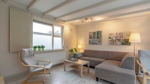 A cozy living room with a grey sofa, a wooden chair, a small table, and a window with potted plants. - Bungalow in Sint Maartenszee Near Beach (Sint Maartensvlotbrug)