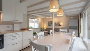 A modern kitchen with white cabinetry, a wooden countertop, and a large dining table with white chairs. There are pendant lights hanging above the table. - Bungalow in Sint Maartenszee Near Beach (Sint Maartensvlotbrug)