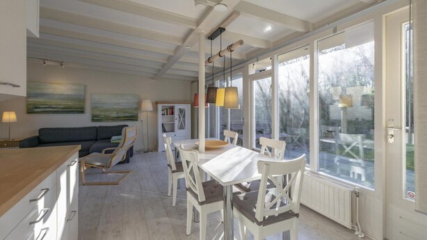 Eine moderne Küche mit weißen Schränken, ein Essbereich mit weißen Stühlen und große Fenster mit Blick ins Freie.