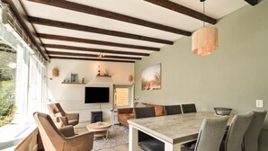 A modern living room with a dining area, wooden beams, a flat-screen TV, and a large window. - Duinland 197 Bungalow in Sint Maartenszee (Sint Maartensvlotbrug)
