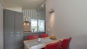 A compact kitchen with grey cabinets, a dining table with red chairs, and a window with striped curtains. - Family Home by the Dunes (Sint Maartensvlotbrug)
