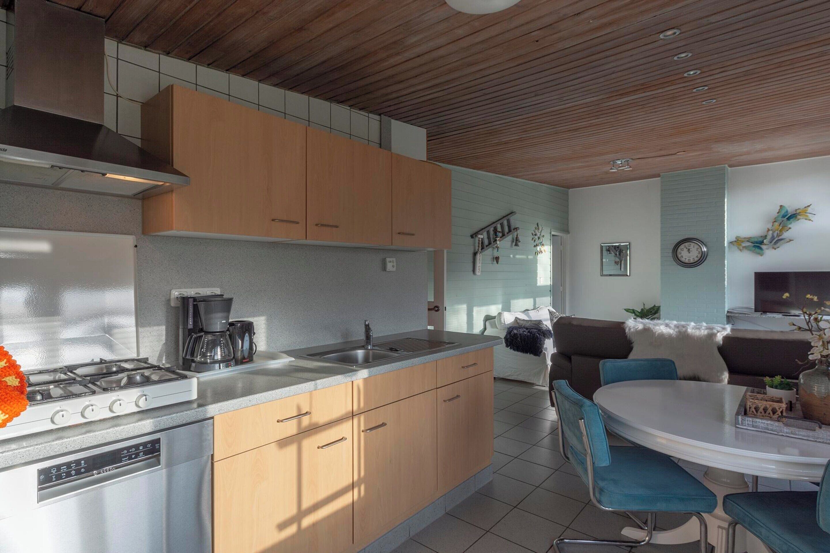A modern kitchen with a gas stove, sink, and wooden cabinets. There is a dining area with a round table and blue chairs.