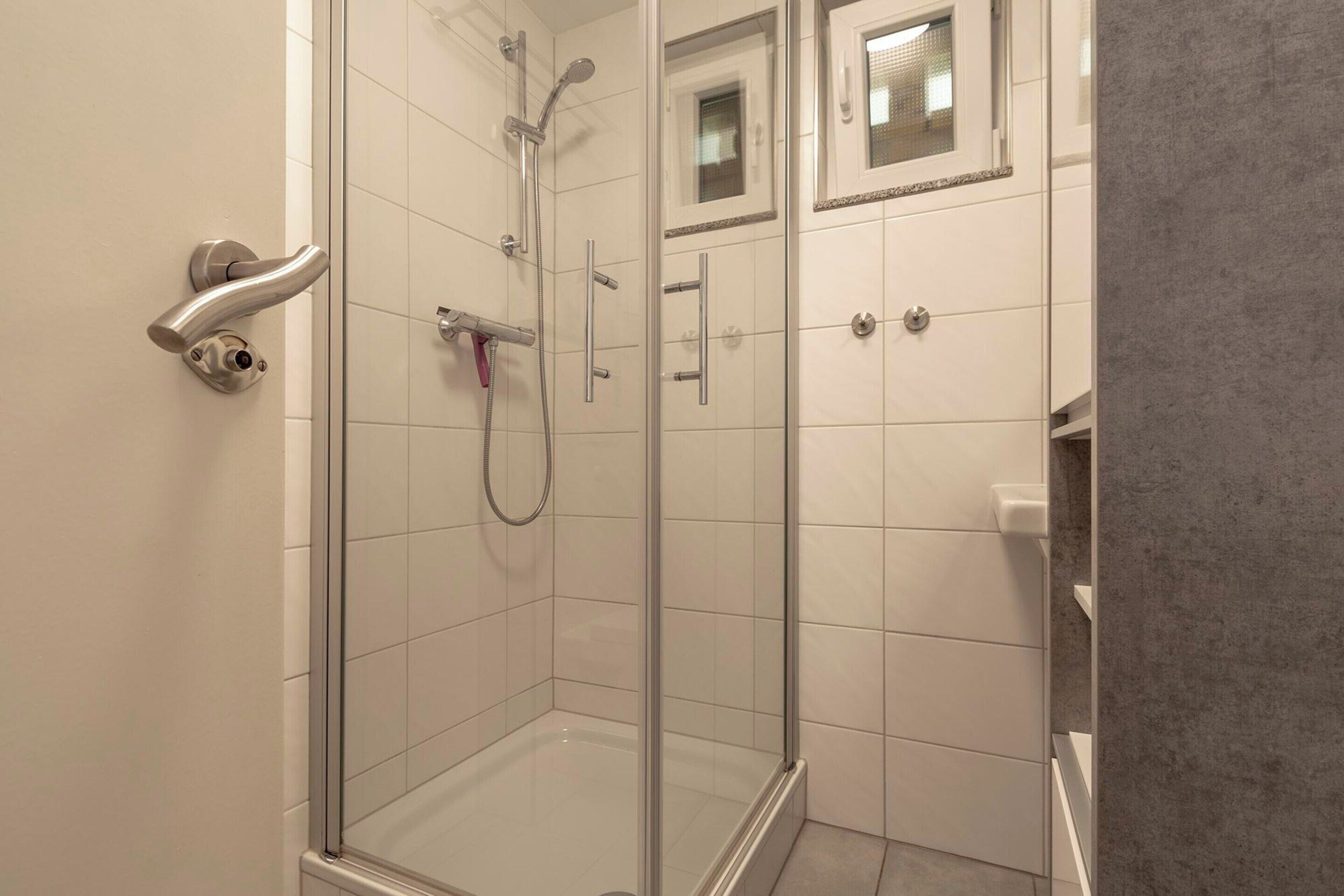 Ein modernes Badezimmer mit Duschkabine aus Glas, weißen Fliesen und einem kleinen Spiegel über dem Waschbecken.