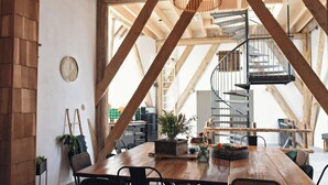 Un comedor rústico con una mesa de madera, sillas y una lámpara colgante. Se ve una escalera de caracol al fondo.