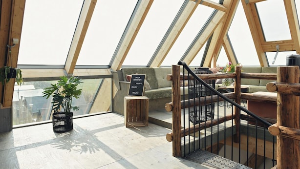 Una habitación con techos altos y techo inclinado, vigas de madera, una escalera, una zona de estar con cojines y una pequeña mesa con una planta.