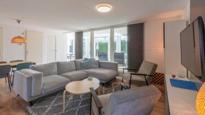 A modern living room with a grey sectional sofa, a round coffee table, and a TV on a stand. There is a dining area with a table and chairs, and a large window providing a view of the outdoors. - Bungalow With Sauna and Pool in Quiet Location (Sint Maartensvlotbrug)