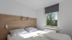 A bedroom with a wooden headboard, a bed with white bedding, two wall-mounted light fixtures, and a window with a view of greenery. - Bungalow With Sauna and Pool in Quiet Location (Sint Maartensvlotbrug)