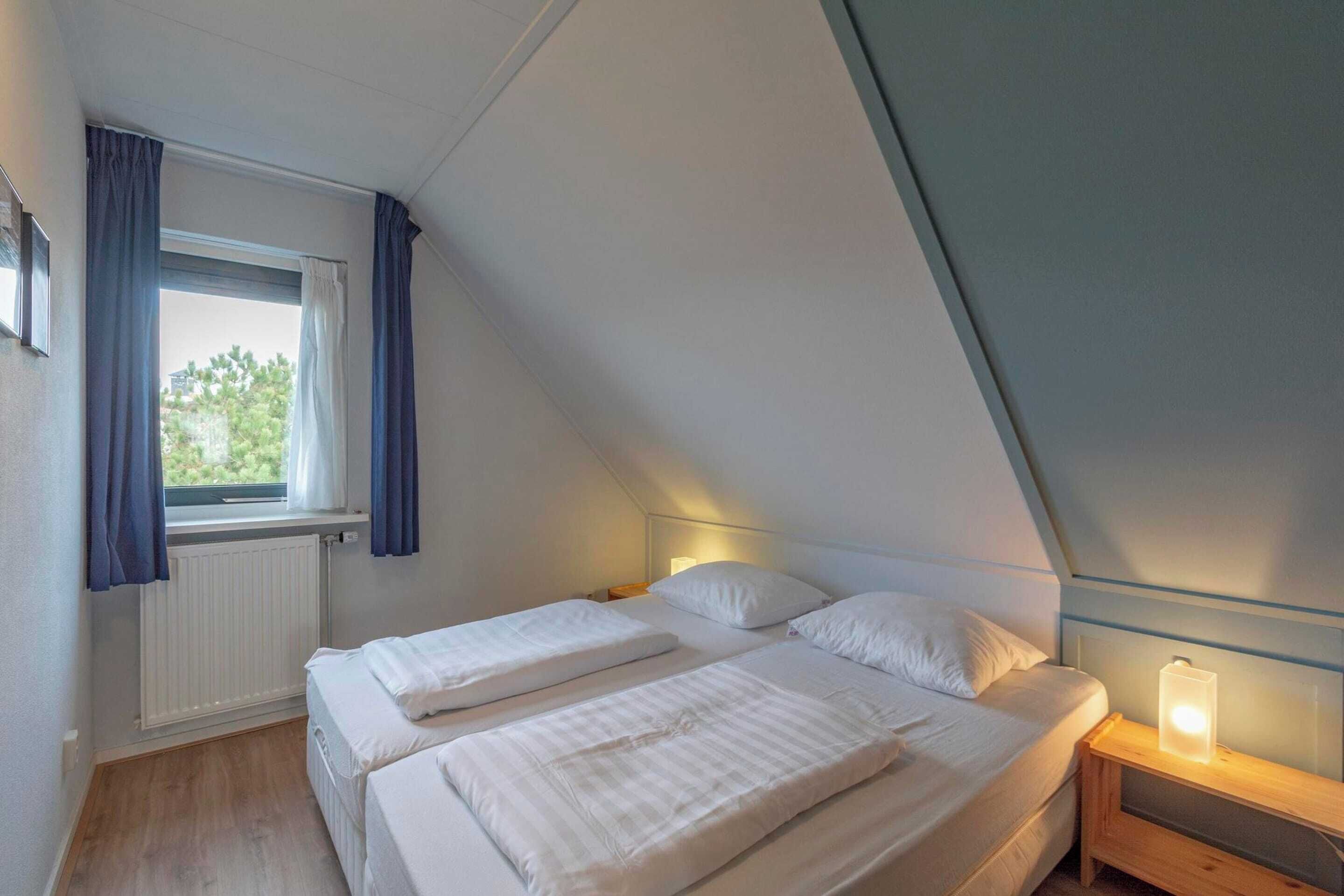 A bedroom with two beds, a window with curtains, a bedside table with a lamp, and a radiator.