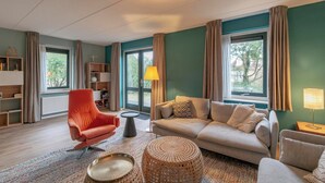 Una sala de estar moderna con un sofá, un sillón y una mesita de centro. Hay una ventana con cortinas, una lámpara y una estantería.