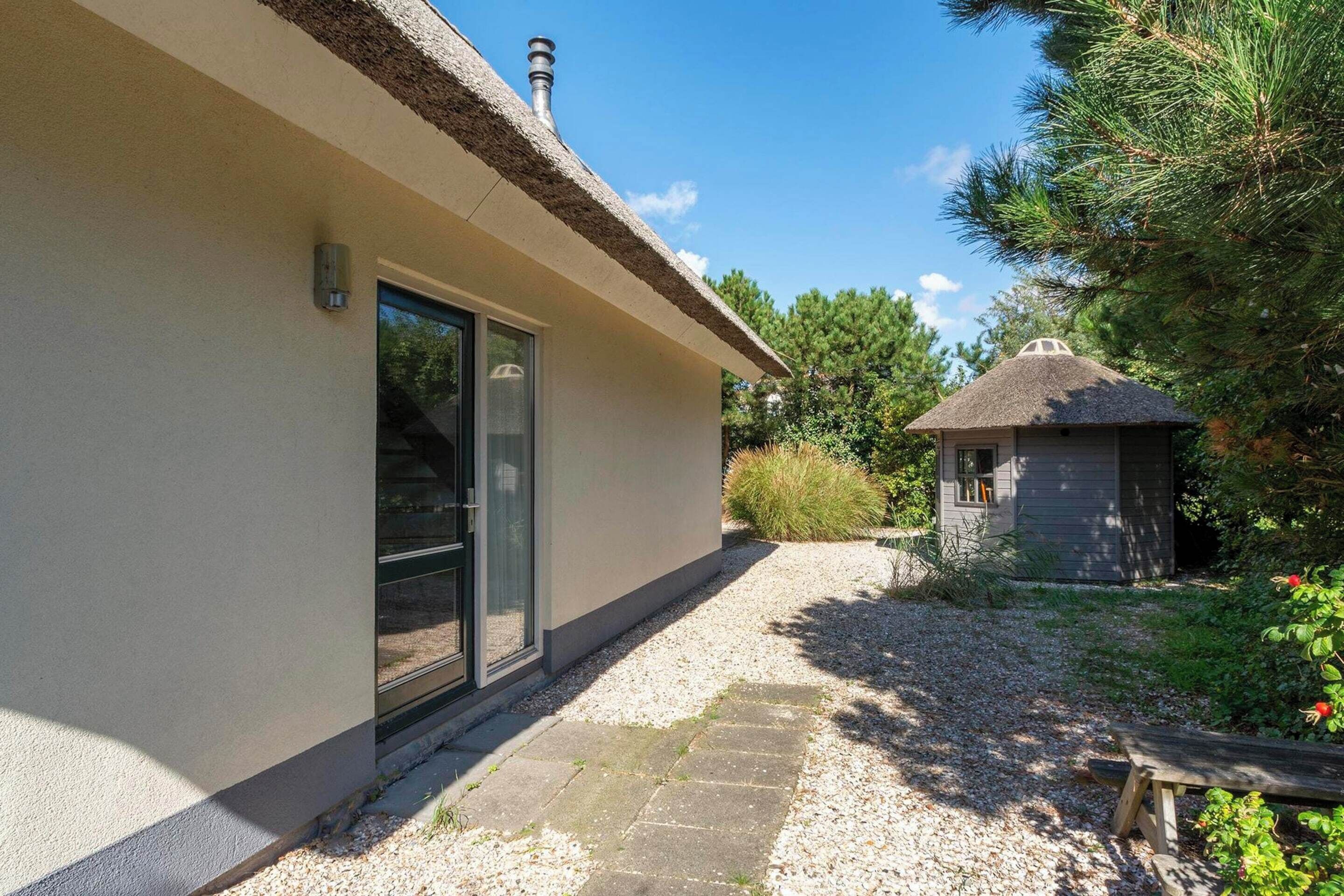 Una casa moderna con techo de paja, un camino de piedra y una pequeña construcción de madera en el jardín.
