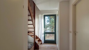 Un pasillo con una escalera de madera, una puerta con marco verde y vistas a árboles afuera.