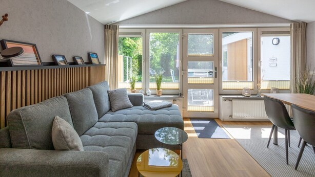 A modern living room with a grey sectional sofa, a small round coffee table, and a dining area with a wooden table and chairs. - Garnekuul 49 Holiday Home in Callanstoog (Callantsoog)