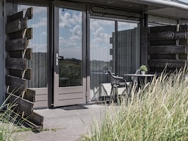 A modern building with a glass door entrance, a small outdoor seating area with a table and chairs, and a view of the surrounding landscape.