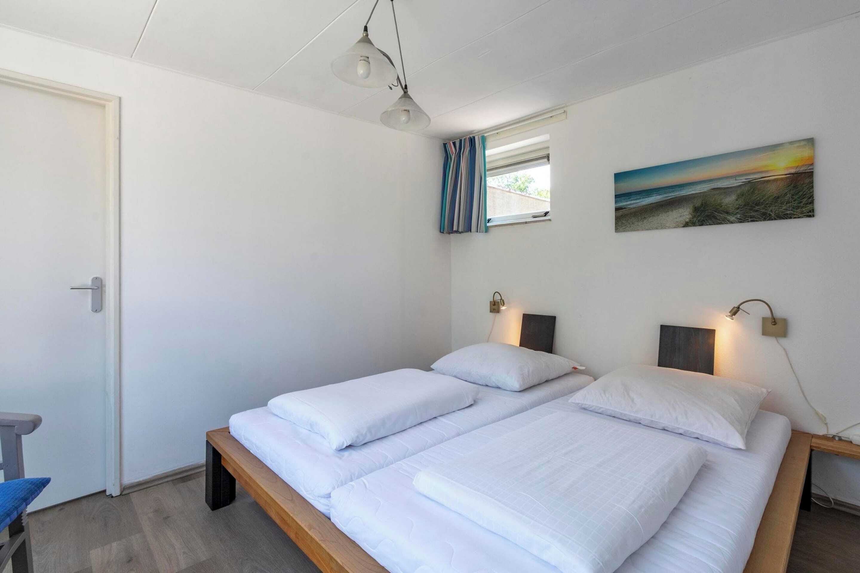 A room with two single beds, a window with curtains, a painting on the wall, and a desk with a lamp.