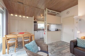 Une cuisine compacte avec des meubles blancs, une table à manger avec des chaises et un fauteuil en osier doté d’un coussin.