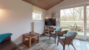 Un salon chaleureux avec un canapé en cuir marron, des fauteuils en osier dotés de coussins, une table basse en bois, un téléviseur à écran plat posé sur un meuble et une grande fenêtre offrant une vue sur l’extérieur.