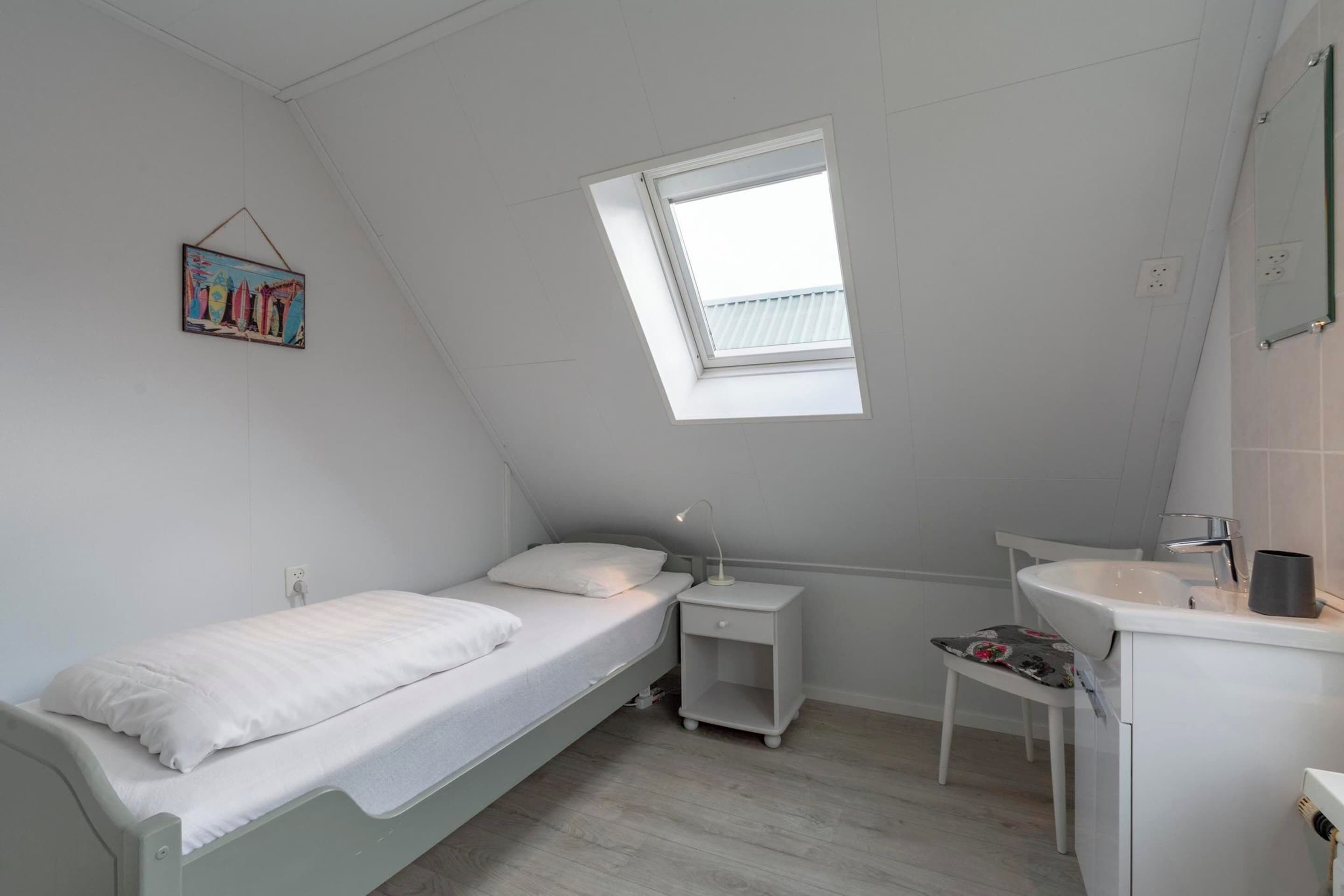 A small, white, single-bed room with a skylight, a bedside table with a lamp, a small chair, and a white sink.
