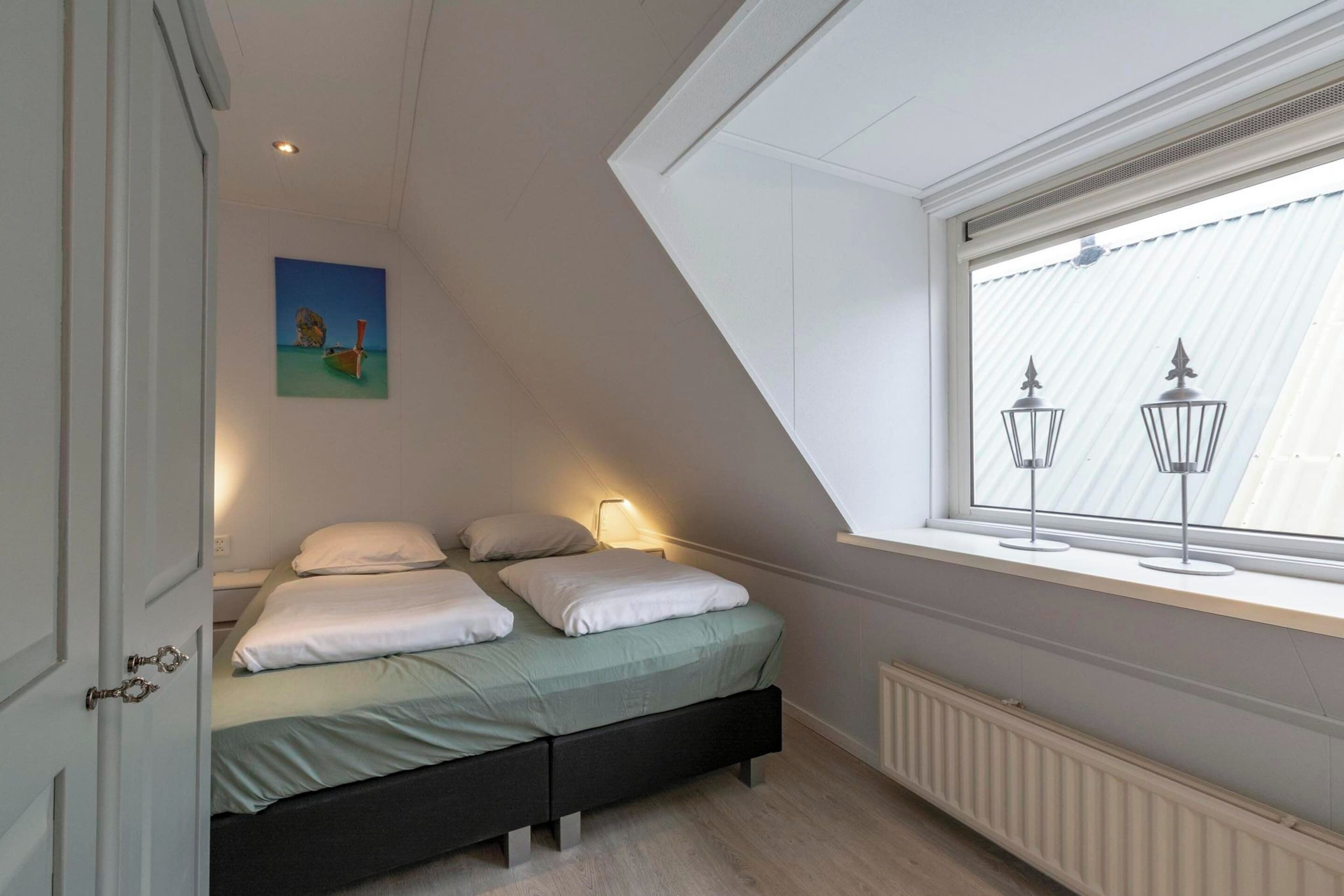 A bedroom with a sloped ceiling, a bed with white bedding, a nightstand with a lamp, and a window with two wall-mounted lights.