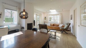 A spacious living area with a wooden dining table, a sofa with cushions, and a large window providing natural light.