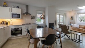 A modern kitchen with white cabinets, a dining table, and chairs. There is a large window with curtains, a sofa, and a TV in the living area.