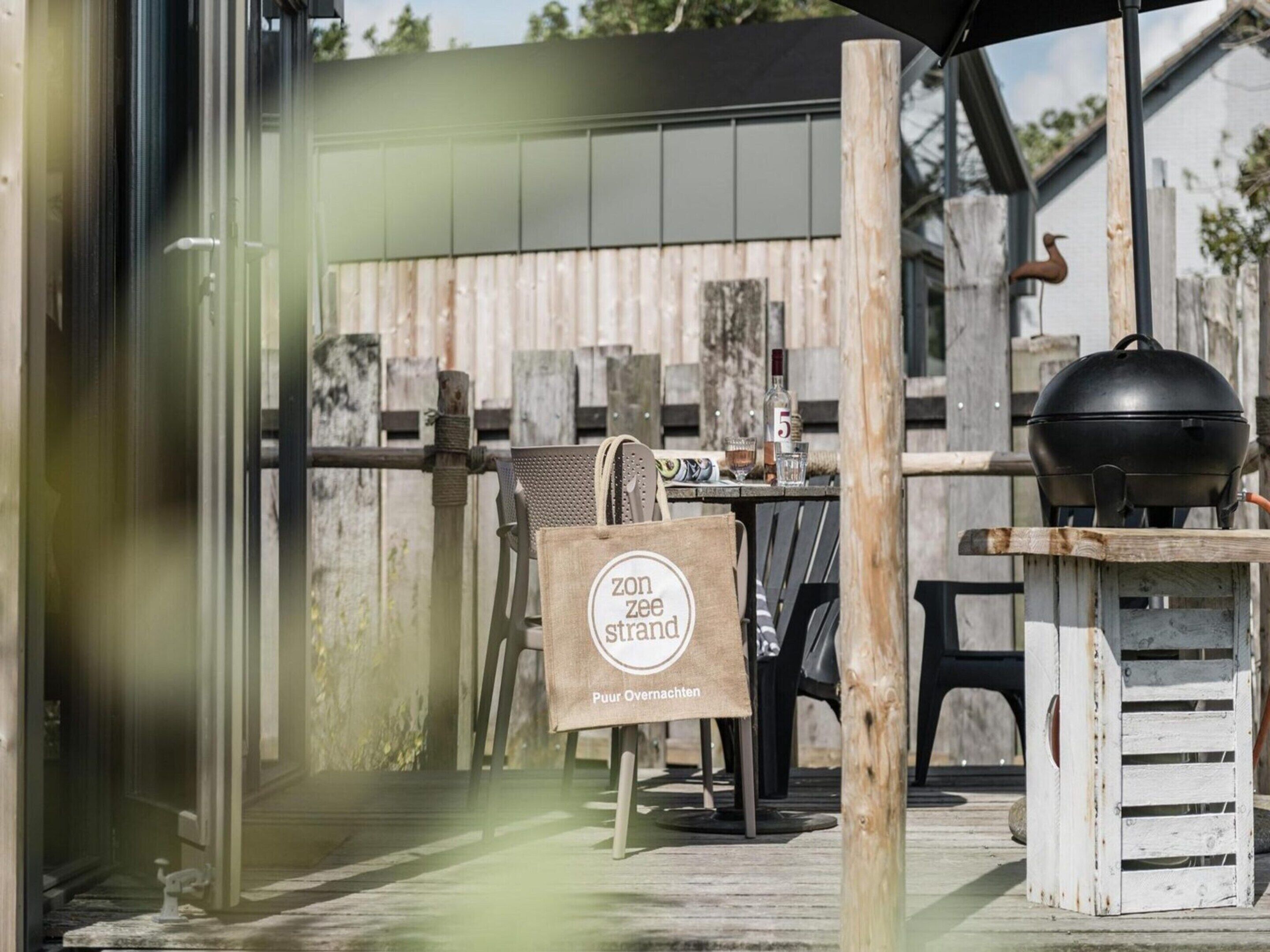 An outdoor seating area with a wooden fence, a barbecue grill, and a table with chairs. A sign with the text "Zon Zee strand" is visible.