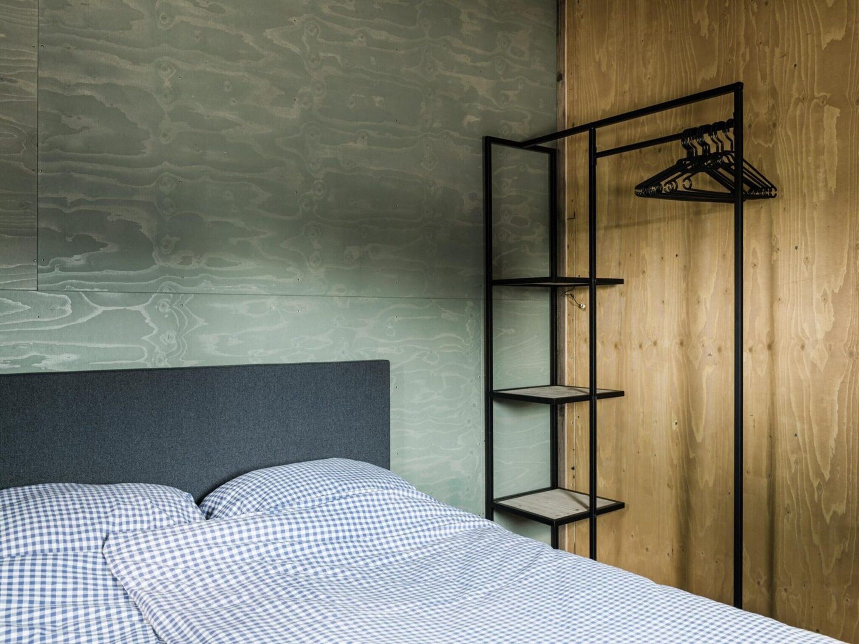 A bedroom with a bed, a grey headboard, a wooden wardrobe, and a black metal shelf unit with a hanger.