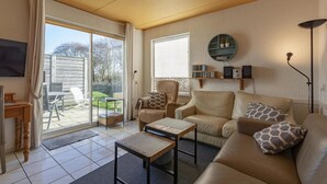A living room with a sofa, armchair, coffee table, and a TV. There is a sliding glass door leading to a patio with outdoor furniture. - Cozy Holiday Home With Terrace (Callantsoog)