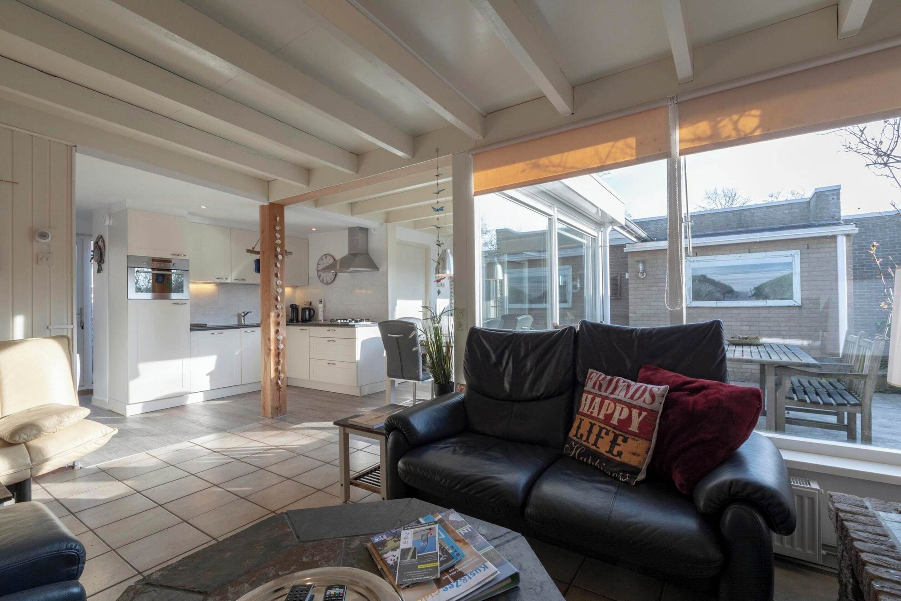 A spacious living room with a black leather sofa, a coffee table with magazines, and a view of the backyard through large windows.