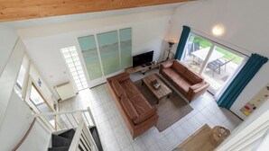 Ein geräumiges Wohnzimmer mit einer großen Couch, einem Couchtisch und einem Fernseher. Eine Treppe führt zu einer anderen Ebene, und ein großes Fenster mit Vorhängen.