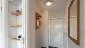 A hallway with white walls, a wooden door, and a mirror with a gold frame. Shelves are mounted on the wall, and a light fixture is visible on the ceiling. - Holiday Home on Car-free Park Garnekuul (Callantsoog)