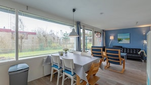 A dining area with a table and chairs, a trash bin, and a window with blinds. - Child-friendly Garnekuul 81 Bungalow (Callantsoog)
