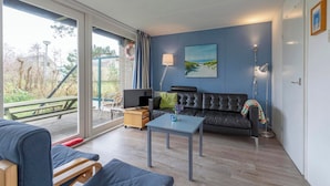 A modern living room with a sofa, a coffee table, and a TV. There is a large window with a view of the outdoors. - Child-friendly Garnekuul 81 Bungalow (Callantsoog)
