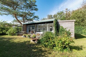 Een modern huis met een glazen uitbouw, een terras met tuinmeubilair en een verzorgde tuin.