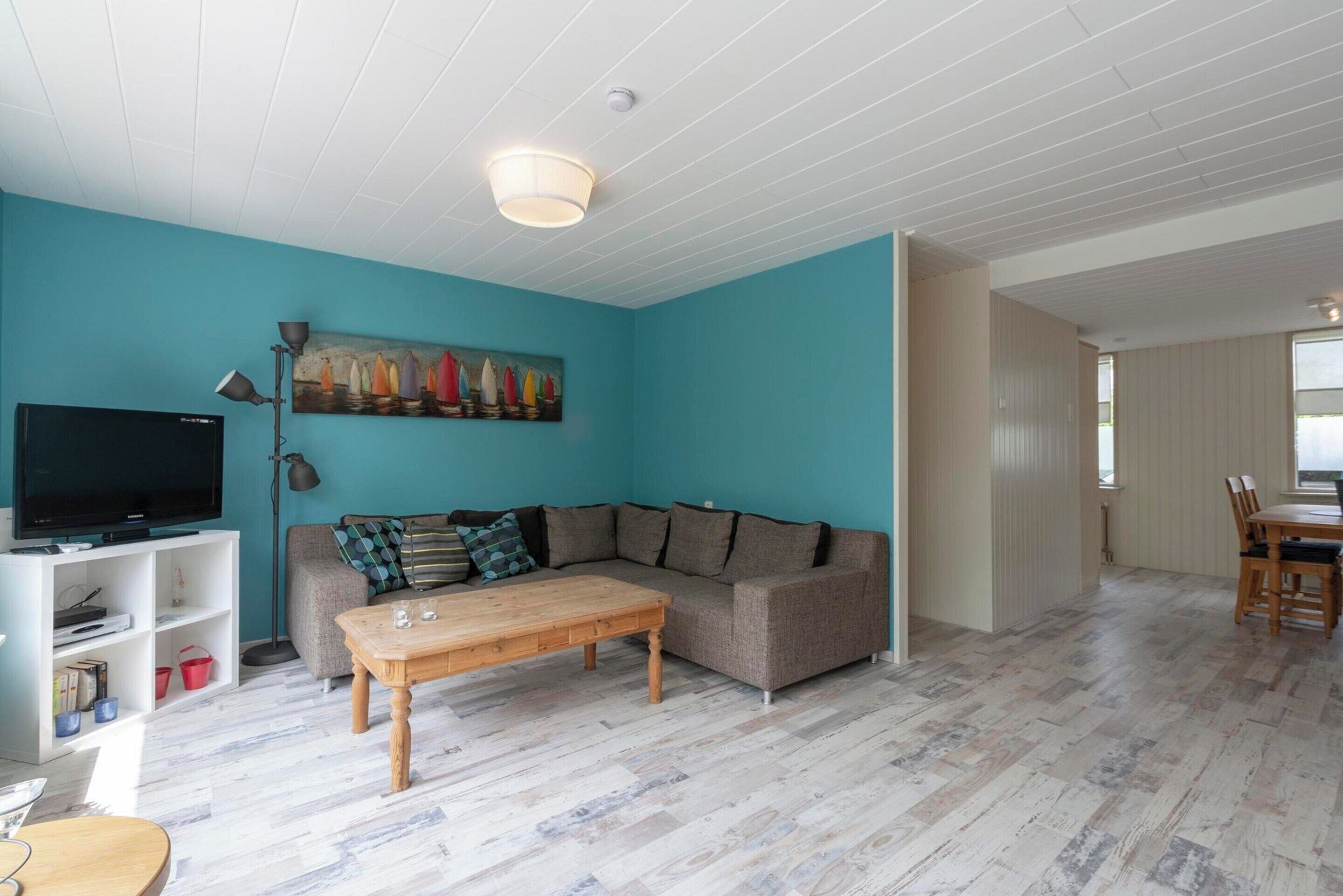 A living room with a teal wall, a wooden coffee table, a grey sofa with cushions, a TV on a white stand, and a painting of sailboats.