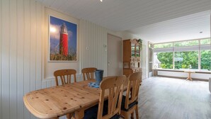 Un coin repas avec une table et des chaises en bois, un tableau représentant un phare et une vue sur la verdure grâce à de grandes fenêtres.