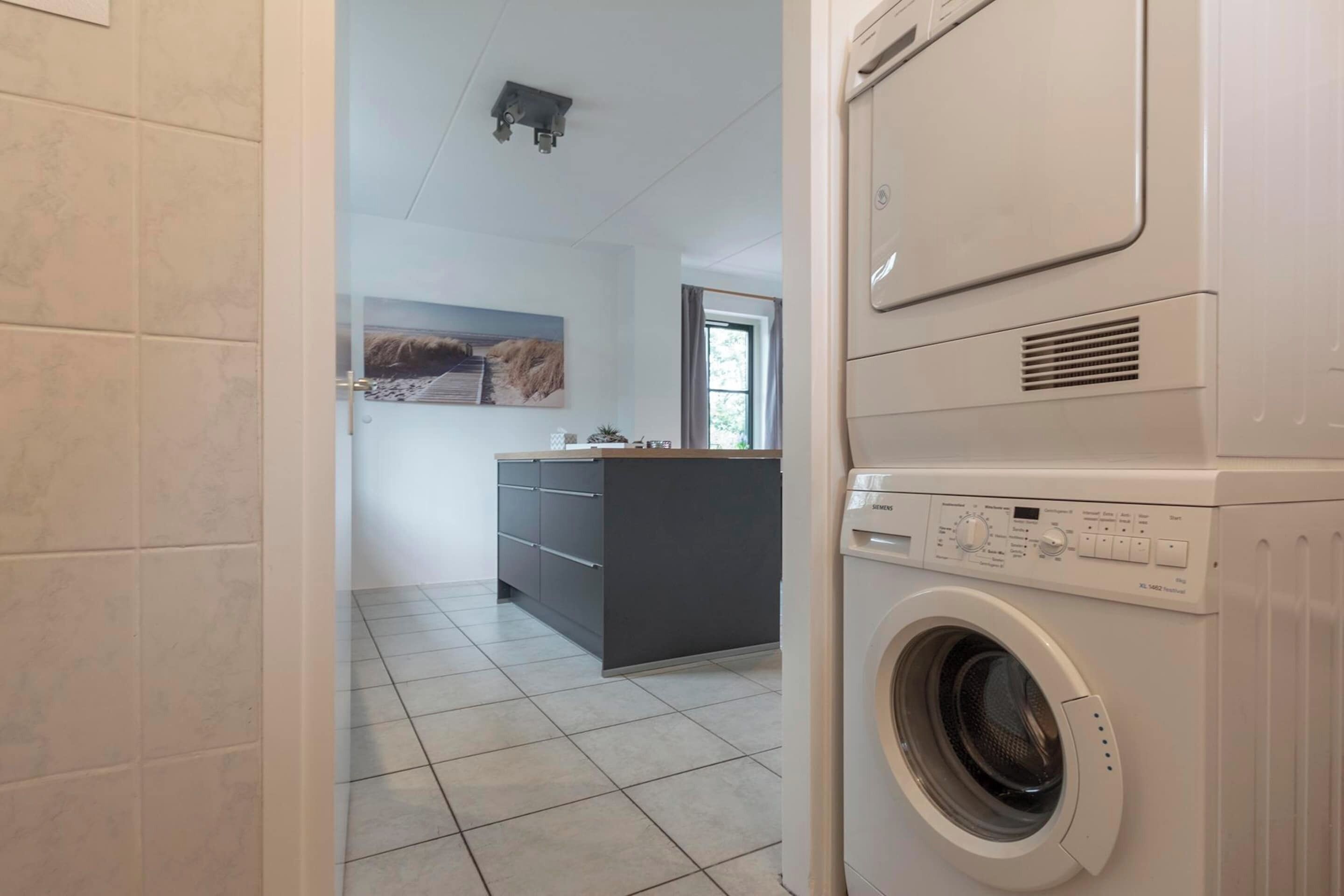 A laundry room with a washing machine and dryer, a tiled floor, and a kitchen area in the background.