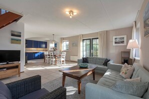 Un vaste espace de vie comprenant un canapé, une table basse en bois et un téléviseur posé sur un meuble en bois. On y trouve également un coin repas avec une table et des chaises, ainsi qu’une cuisine à l’arrière-plan.