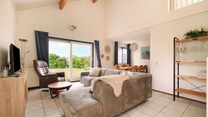 A modern living room with a sofa, a television, a dining area, and a large window. - Villa in Julianadorp Near Sandy Beach (Julianadorp)