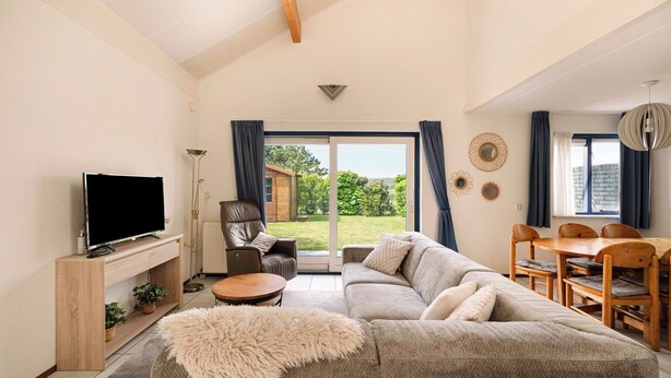 A living room with a sofa, TV, wooden table, and a view through a glass door to the outside. - Villa in Julianadorp Near Sandy Beach (Julianadorp)