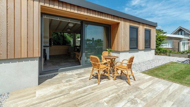 Una casa moderna con fachada de madera, una puerta de vidrio que da a un espacio interior y un patio con mobiliario de mimbre.