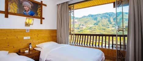 Quarto de hotel com duas camas, cabeceira de madeira, um quadro, mesa de cabeceira, abajur e varanda com vista para o campo.